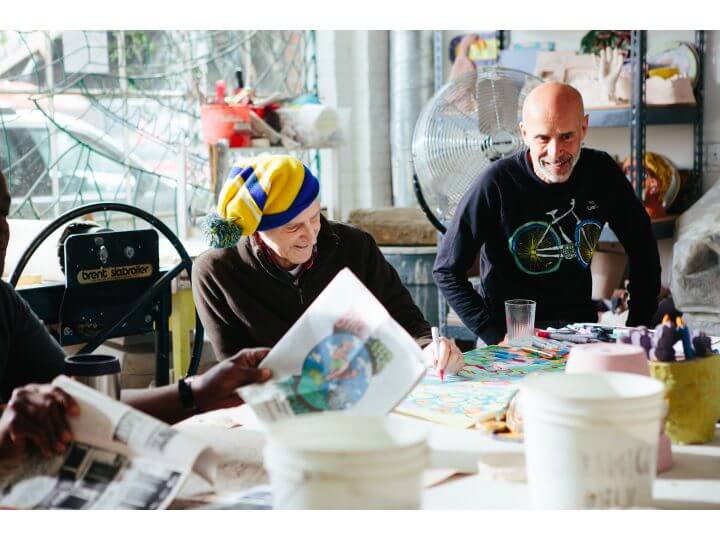 Image description: Two men sit at a work space together. Barry Regan, the one of the left wears a yellow winter hat. To the right Tom di Maria wears a blue longsleeve shirt. 