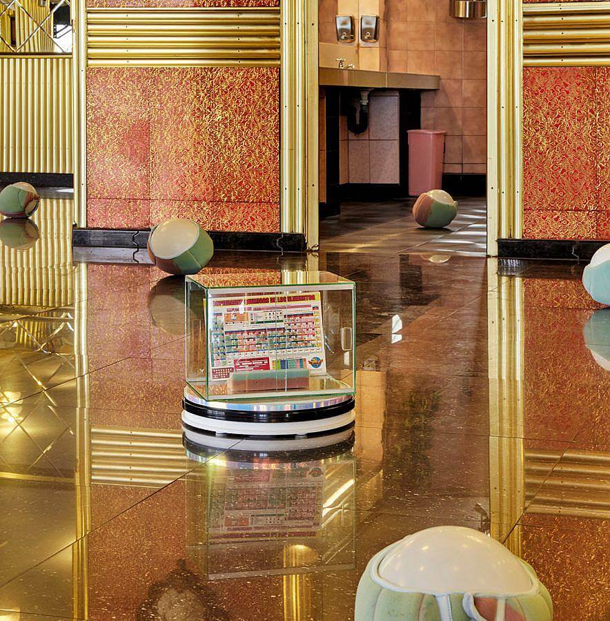 Image description: A display area of. dark marble floor reflects the surroundings. In the center of the photo a case which holds a periodic table of sponge is displayed. Balls wrapped in green and pink sponge are displayed around the room.