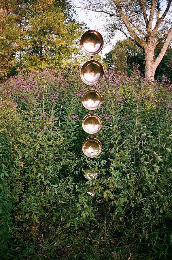 Image Description: Standing amongst a big flowering bush is a tall stack of gold singing bowls. The center of the bowls are facing the viewer.