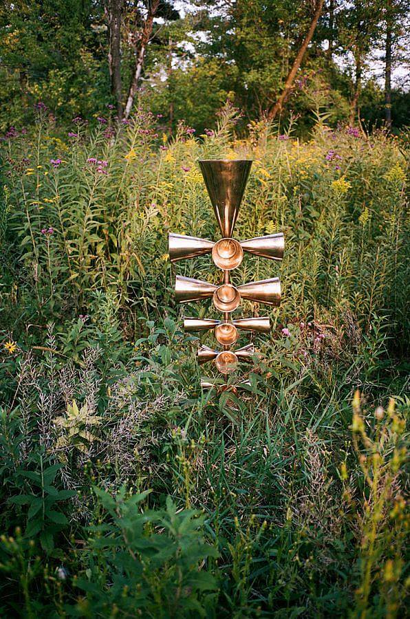 Image Description: Standing amongst a dense mix of wild plants is a stack of golden bells. On each level, there are cone-shaped bells on all four sides and the bells progressively get bigger towards the top. The largest bell is stacked on top.