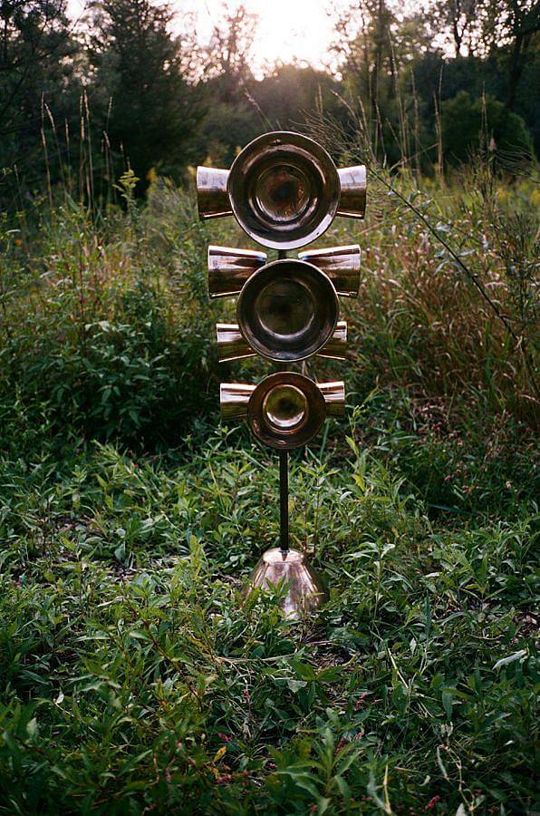 Image Description: Standing amongst various wild plants and streets are a set of wide cup-shaped bells arranged along a standing bowl. Facing the viewer are three wide bells that get progressively taller towards the top. Along each side of this stack are four smaller cylindrical bells.