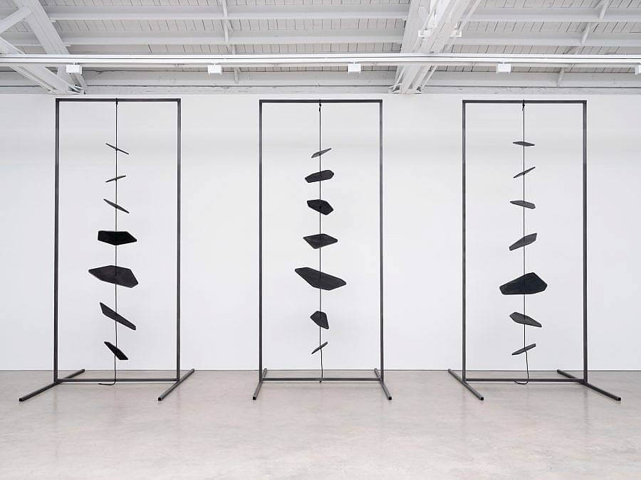 Image Description: Facing the viewer are three metal rectangular free-standing frames. Stretched through the center of each is a black rope with seven spinning black metal shapes of various sizes.