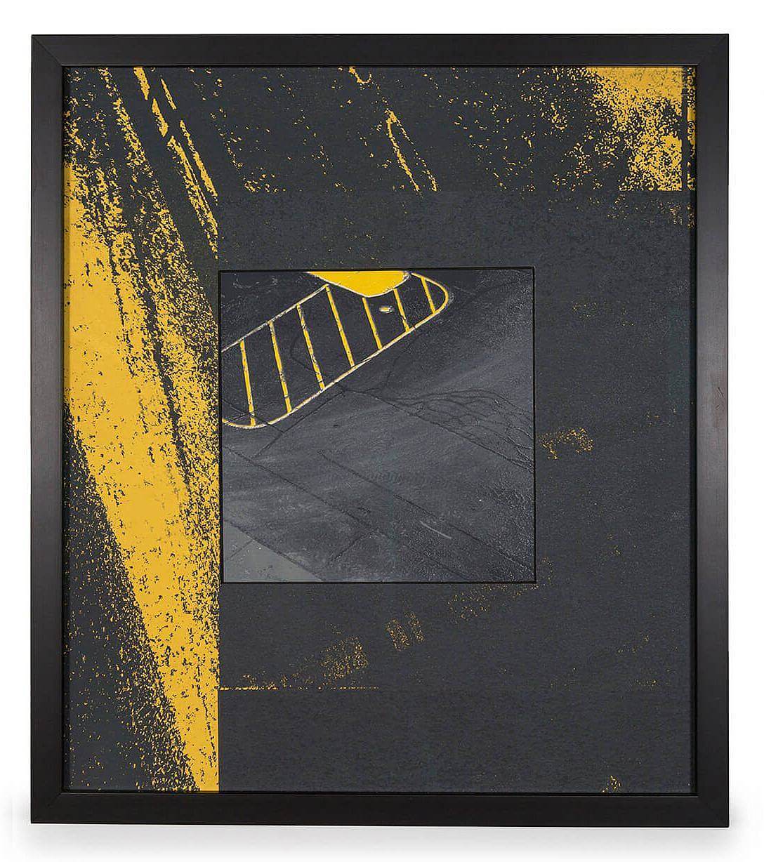 Image Description: There is a thick border around the image depicting a close-up image of black asphalt with yellow markings. The image in the center of the piece is a birds-eye view of an asphalt parking lost with a yellow outline of an oval with diagonal stripes going across. 