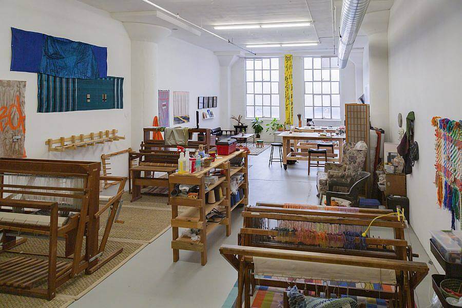 Image Description: This is an image of a communal studio space with multiple looms and other tools for fiber work. There are large windows at the far end of the room and various weavings hanging on the wall.