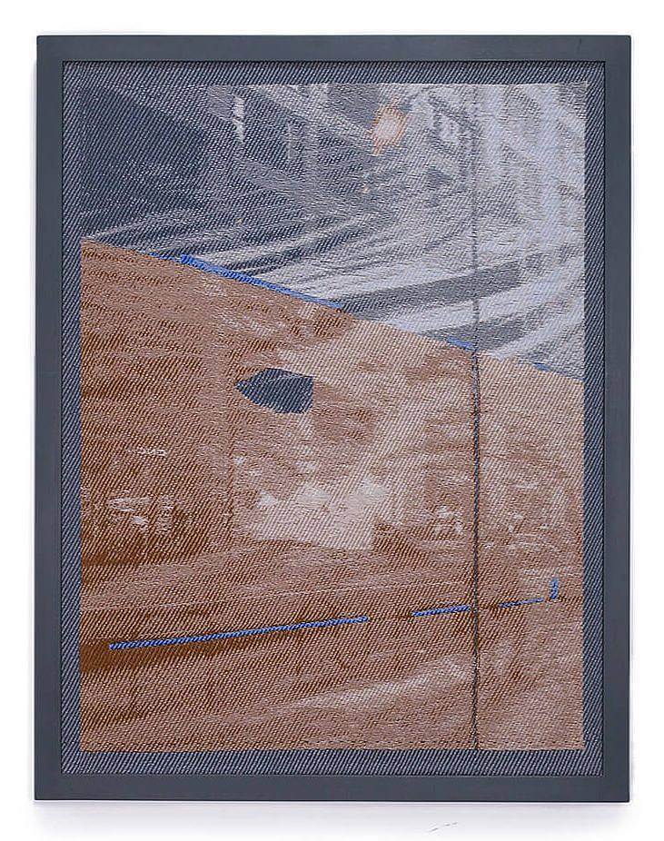 Image Description: This is a vertical rectangular weaving in a dark blue-grey frame. The weaving depicts a street scene with a tall office building, bike racks, and a street lamp. The top of the image is down in shades of grey and the bottom is in variations of an orange-brown.