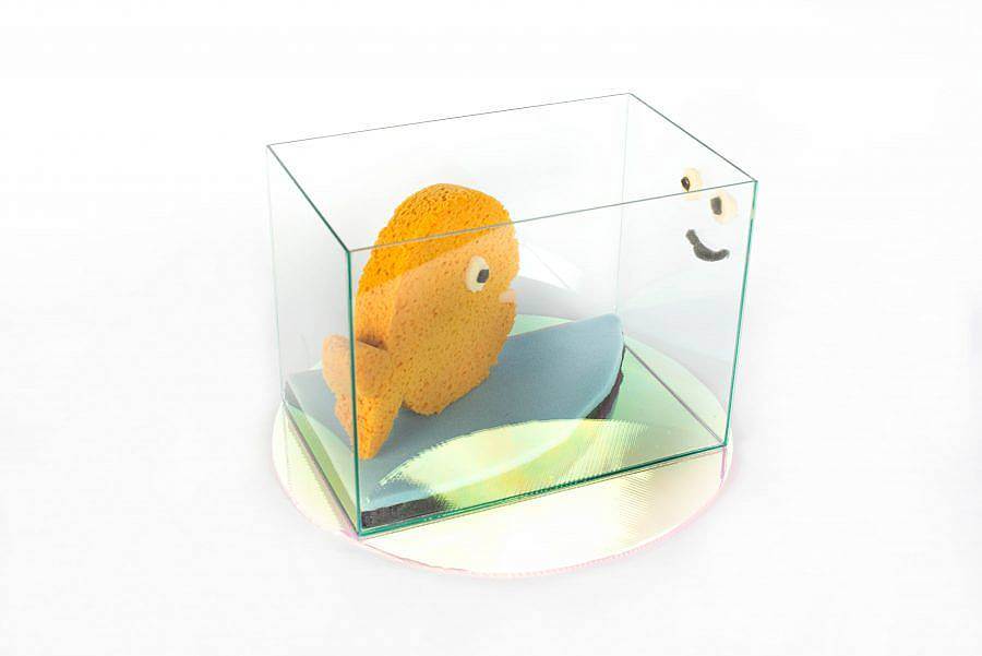 Image description: A fish out of sponge is in a glass case. A small sponge smiley face is on the side of the case opposite of the fish. 