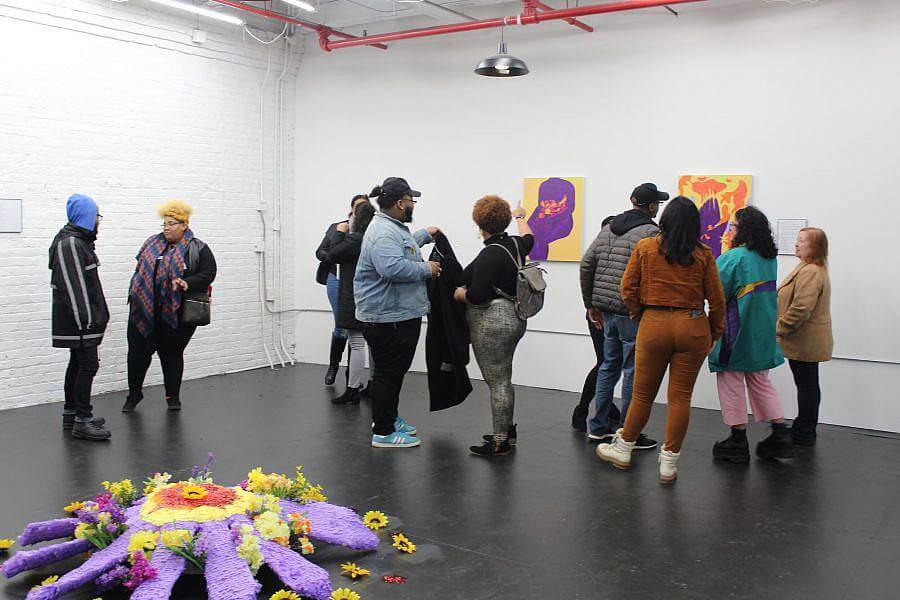 Image description: A group of eleven people are in an exhibition space. To the left a large purple and yellow pinata flower is placed on the ground. 