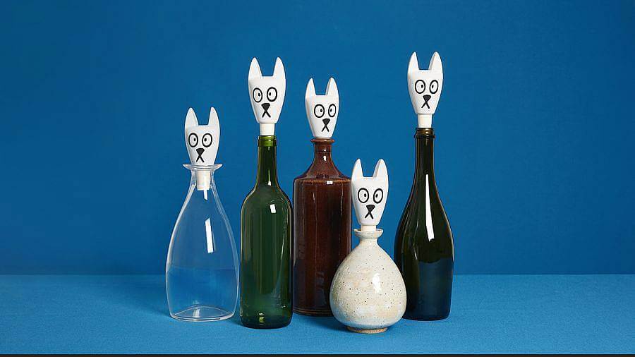 Image description: A series of five vessels are displayed in front of a blue background. The vessels are all glass, except for the fourth from the left which is ceramic. They all have white cat head bottle stoppers in them. The bottle stopper is fit with pointy cat ears, big eyes, and a small mouth. 