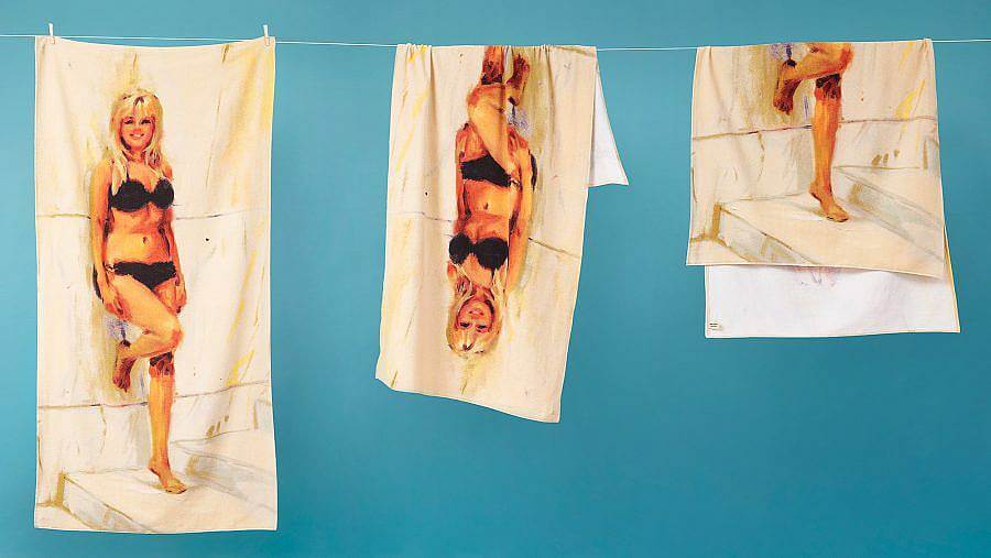 Image description: A series of three towels hang on a line in front of a yellow blue background. The towels feature an illustration of a blonde woman leaning up against a stone wall wearing a black bikini.