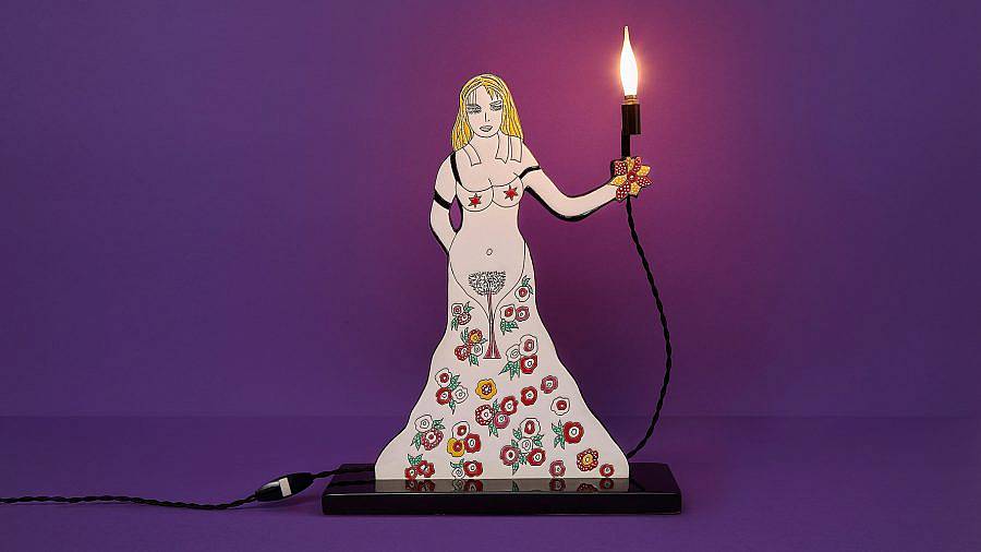 Image description: A light fixture is displayed against a purple background. An illustrated white woman is naked with red stars on her nieepls and an illustrated tree to cover her pelvic region. Flowers lay out where her legs should be forming a skirt around the bottom half of her body. She holds a lamp, where a real life fixture illuminates the setting. 