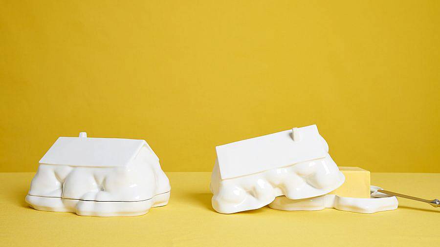 Image description: Two butter dishes are placed on a mustard yellow table in front of a similarly colored background. The butter dishes are shaped like homes that have natural formations have grown around them. The bottom half of their forms appears organic. The right butter dish is left partially ajar to display a stick of butter sticking out. 