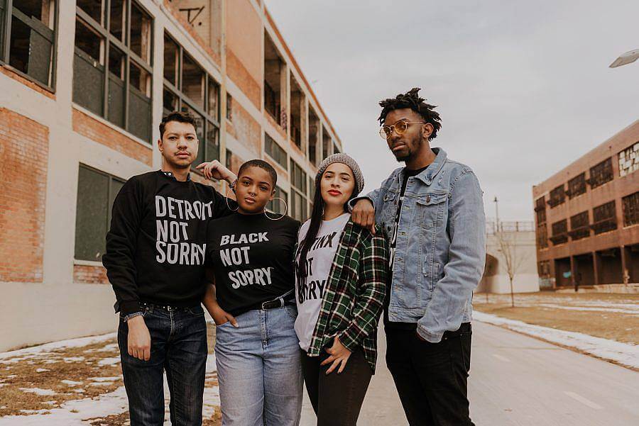 Image Description: There are four people standing together in front of a set of buildings facing the camera. Three are wearing Not Sorry Goods shirts that say "Detroit Not Sorry", "Black Not Sorry", and "Latinx Not Sorry". 