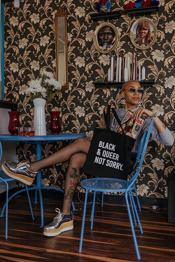 Image Descriptions: There is a figure sitting in a blue metal chain pulling a magazine out a black tote bag. The tote bag says Black & Queer Not Sorry on it and the person is wearing sunglasses and has their legs crossed. There is a floral black and gold wall-paper in the background.