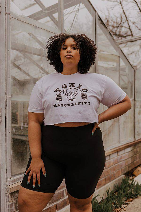 Image Description: The model is standing with one hand on their hip facing the camera in front of a green house. They are wearing black bicycle shorts and a white crop top that says "Toxic Masculinity". There is also an illustration of a man lifting weights on the shirt.