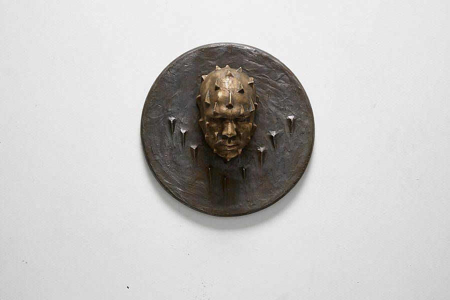 Image description: This is a circular wall piece with a face protruding from the center. The face is looking down and is covered with spikes. There are spikes on the plaque in a "V" formation under the face as well.