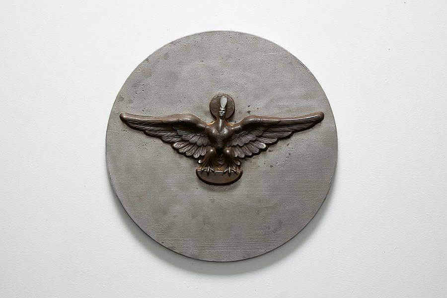 Image Description: This is a circular wall piece with a bird with a small human head embossed in the center. The wings are outstretched and the bird is perched on a half oval. There is a small circle around the human head and the head is wearing a tall narrow hat.