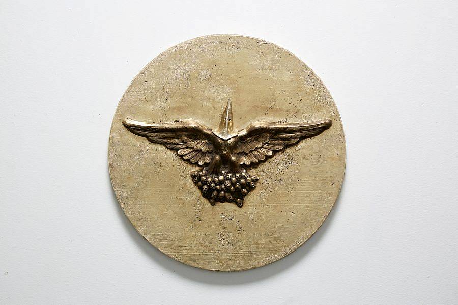 Image Description: This is a circular wall piece with a figure with outstretched bird wings embossed into the center. The man appears to be standing on a mass of small balls and is wearing a Ku Klux Klan hood over his head. 