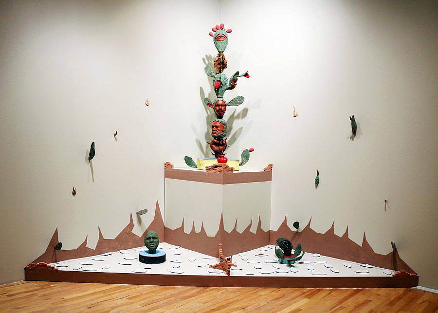 Image Description: This is an installation centered in the corner of a room. On a triangular pedestal, there is a wall cacti with red faces on the leaves. Surrounding it, on a slightly raised platform near the floor, are two green faces in cactus forms and intersecting sets of terracotta stairs.