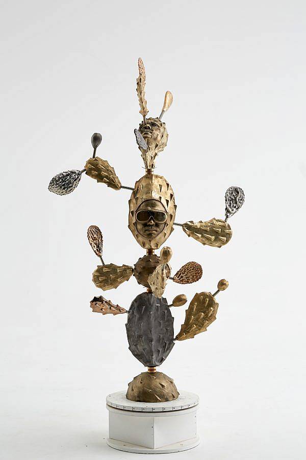 Image Description: This is a metal sculpture of a cactus in metallic gold and silver tones. There are two faces protruding from the cactus. The face facing the viewer is wearing aviator sunglasses and has a mustache. 