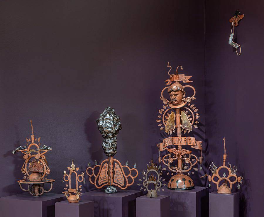 Image Description: This a collection of ceramic pieces on pedestals in a dark purple room. The pieces of varying heights and a visually complex, incorporating loops, lungs, leaves, flames, and more. They are all a terracotta orange with touches of black and a metallic dark green.