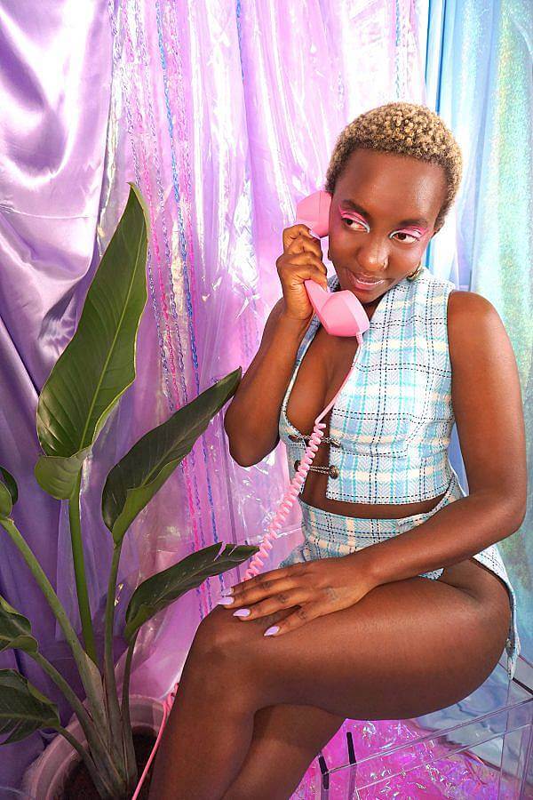 Image description: A dark-skinned black woman with short blonde hair wears a baby blue plaid two piece. The top is a vest that ties in the middle and the bottom is straight mini skirt with a slit down the leg. She holds a pink phone in front of a satin purple backdrop. A bird of paradise is next to her. She is sitting on an a clear acrylic stool.