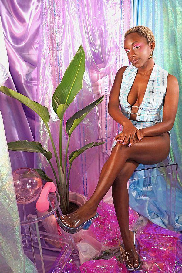 Image description: A dark-skinned black woman with short blonde hair wears a baby blue plaid two piece. The top is a vest that ties in the middle and the bottom is straight mini skirt with a slit down the leg. A pink phone sits on an acrylic table in front of a satin purple backdrop. A bird of paradise is next to her. She is sitting on a clear acrylic stool.