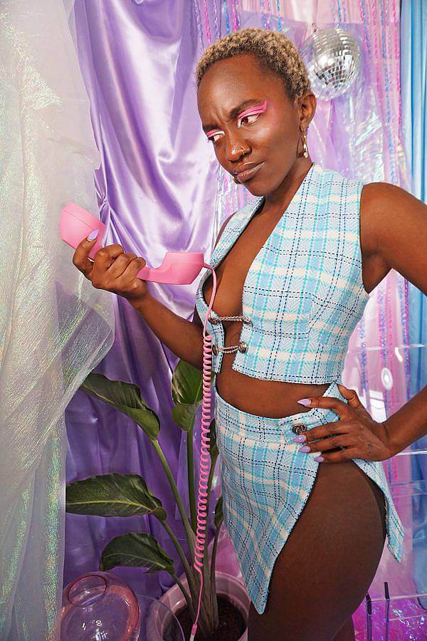 Image description: A dark-skinned black woman with short blonde hair wears a baby blue plaid two piece. The top is a vest that ties in the middle and the bottom is straight mini skirt with a slit down the leg. She holds a pink phone in front of a satin purple backdrop. A bird of paradise is next to her. She holds the phone out from her and looks at it with playful and doubtful face.