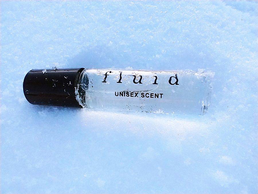 Image Description: This is an image of a small perfume bottle with a black lid laying in the snow. "Fluid. Unisex Scent." is written on the side of the bottle.