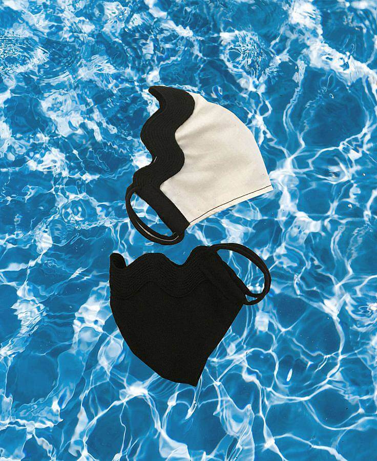Image Description: This is a photograph of two face masks on top of a blue water background. The top of both masks is lined with thick wavy rick-rack. The mask on top of white with black rick-rack and the mask on the bottom is all in black.