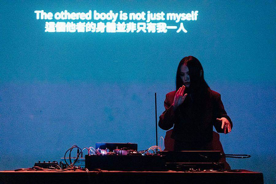 Image description: In this photograph Yilei performs live at Rich Mix. In the photo they are standing behind some electronic music equipment and behind them the words "The bothered body is not just myself" are projected in white onto a deep blue backdrop. Yilei is illuminated in red light.