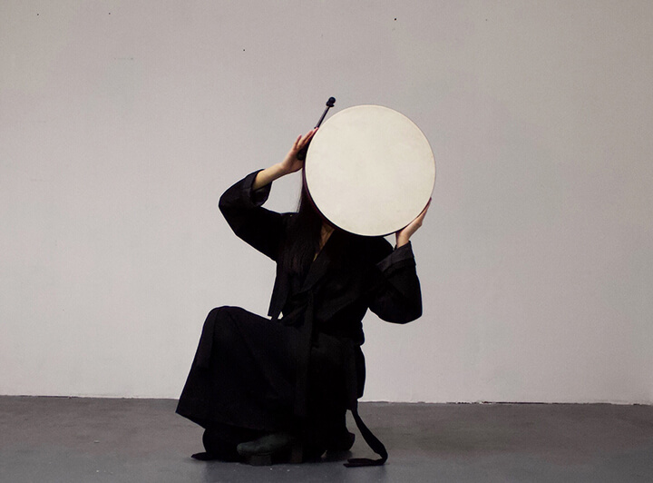 Image description: This image is a photograph of Li Yilei. They are pictured squatting in a room with white walls and concrete floors. They are holding a large, round, beige drum in front of their face. They are dressed in all black.