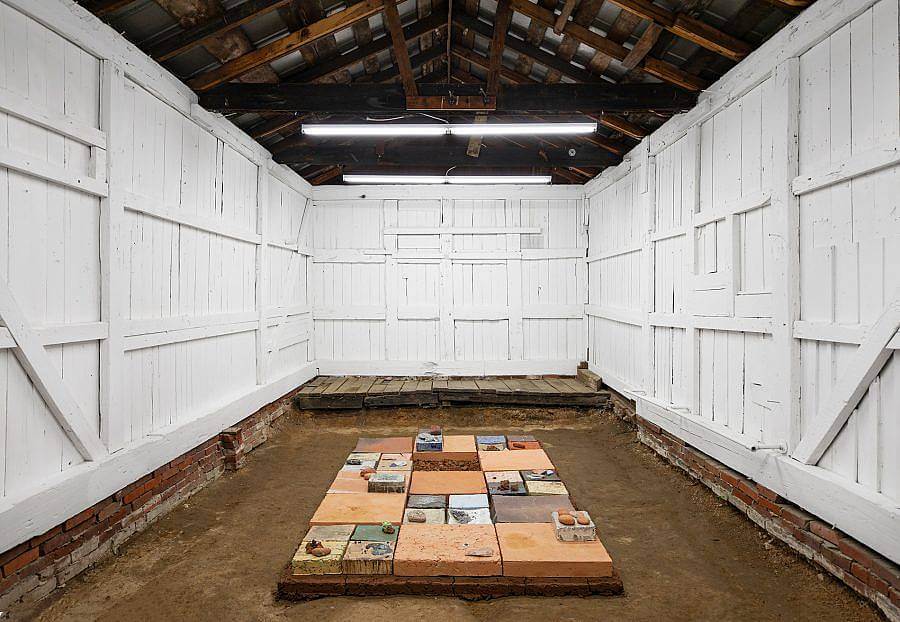 Image Description: This is an installation shot of Bad Water's most recent exhibition. On the floor is a large rectangular platform with various square tiles made of different types of clay layered on top. There are also various stones of different colors and sizes scattered on top. This sculpture is in a small room with white wooden walls reminiscent of a barn interior.