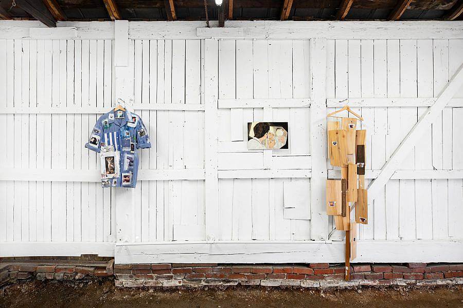 Image Description: Two objects on wooden hangers hang against the white wooden walls of the gallery. One is a blue button-up short-sleeved shirt with photographs attached all over. The other hanging object is a set of thin wooden sheets of different heights, lengths, and widths. In between both hanging object is a small painting of a figure looking in a mirror.