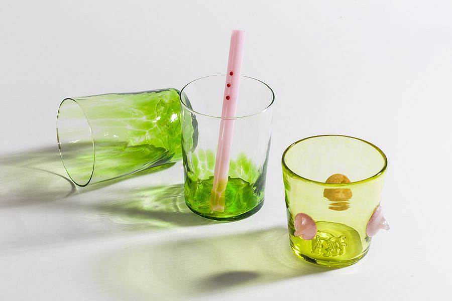 Image Description: There are three glasses in the photograph; the two to the left are transparent with bright green speckled on the bottom. The cup to the right is a more yellow-toned green with three light pink three dimensional dots along the side. There is a pink glass straw in the center cup with three red three dimensional dots lined up at the top. 