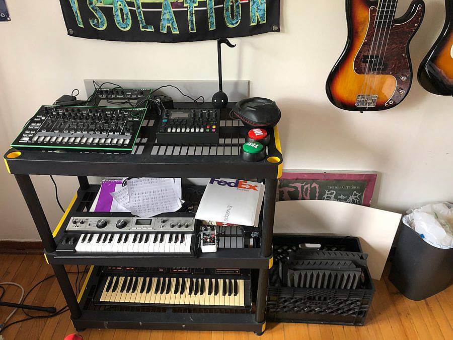 image description: This image features Forget Basement's workspace and musical equipment. There is a table with several panels of knobs and switches, below there are various keyboards accompanied by papers and wires. On the wall there is a poster with the work "isolation" printed in green next to two guitars hanging on the wall.