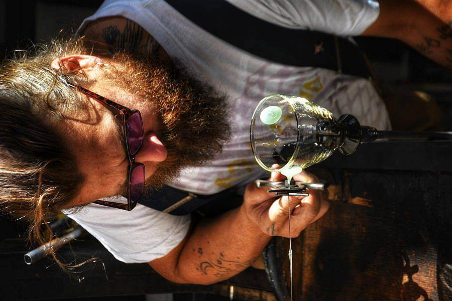 Image Description: This is a photographed of founder Eli working on a glass. He is leaning down to cut a thin stream of glass that form a three dimensional dot on a cup. He is wearing sunglasses.