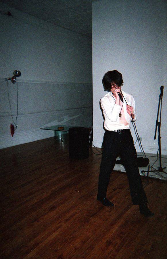 image description: This is a photograph of Forget Basement performing. They are in a room with dark wood floors and white walls. The photo is taken with a flash in an otherwise dark room. They are singing into a microphone that they are holding with their right hand and in their left hand they are holding the cable. They are wearing an open, white, button-up shirt, black slacks, and black dress shoes.