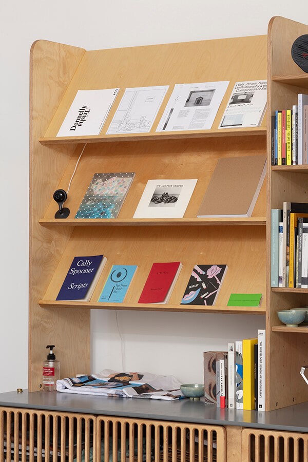 image description: This photograph shows the current book display at the Fulcrum Press. There are twelve books of varying sizes and colors displayed on a wooden book shelf with three wide, slightly slanted shelves to exhibit the front covers of the books.