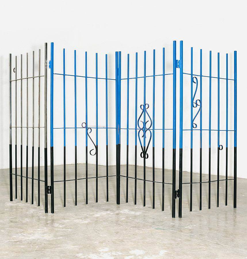 Image Description: This is a large free-standing sheet of wrought iron fencing. It is set up in a zig zag formation and there are various common fencing symbols placed throughout the piece. The top of the piece is painted blue and grey with the bottom painted black.