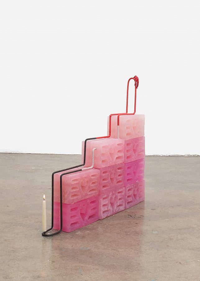 Image Description: There are a set of nine breeze blocks cast in pink soap assembled like a staircase. Steel rebar runs over and through the blocks stacked on top holding everything together. There is a lit tapered candle balanced on the rebar at the bottom of the staircase and there are hearts in the design of the breeze blocks.