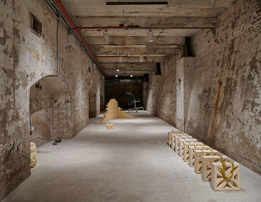 Image Description: This is an installation in a large basement hallway. There is a set of tan breeze blocks casted in cocoa butter lined up with steel rebar along the top. In the distanced is a formation of cocoa butter bricks arranged in a corner formation.