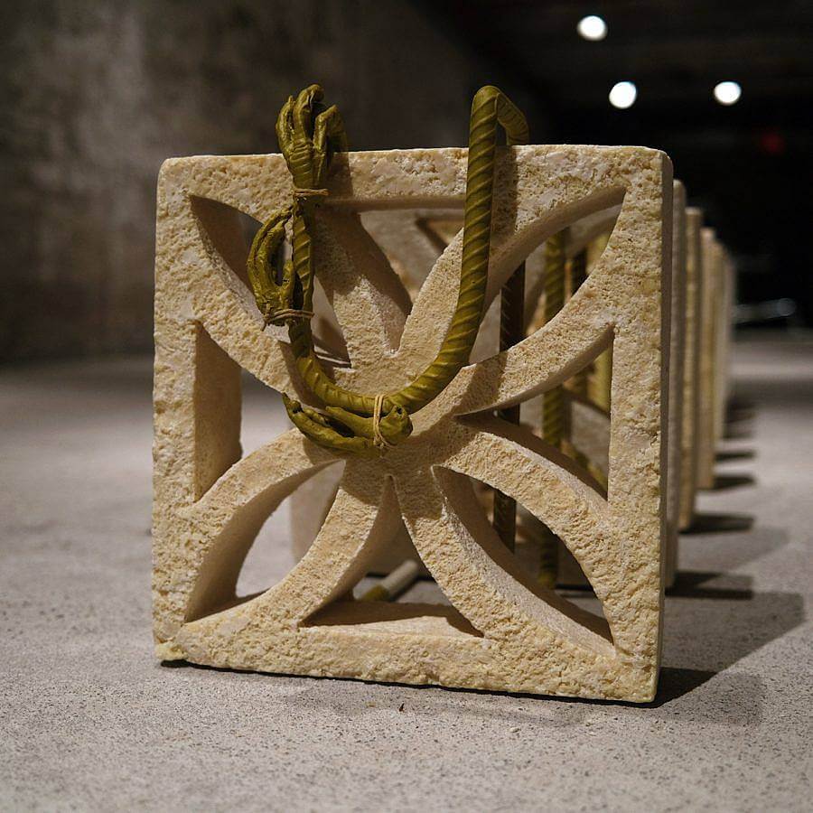 Image Description: This is a square breeze block cast in tan cocoa butter. There is steel rebar in a "U" attached to the top with steel-cast chicken feet tied to it. This breeze block is set up in a long row of others identical to it.