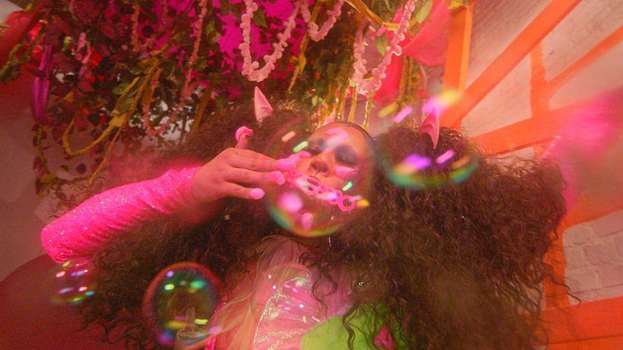 Image Description: This is a still of a person with big curly pigtails blowing bubbles. They have big pink globs on their fingernails and are wearing a pink and green outfit. There is a large plant with streamers in it above them.