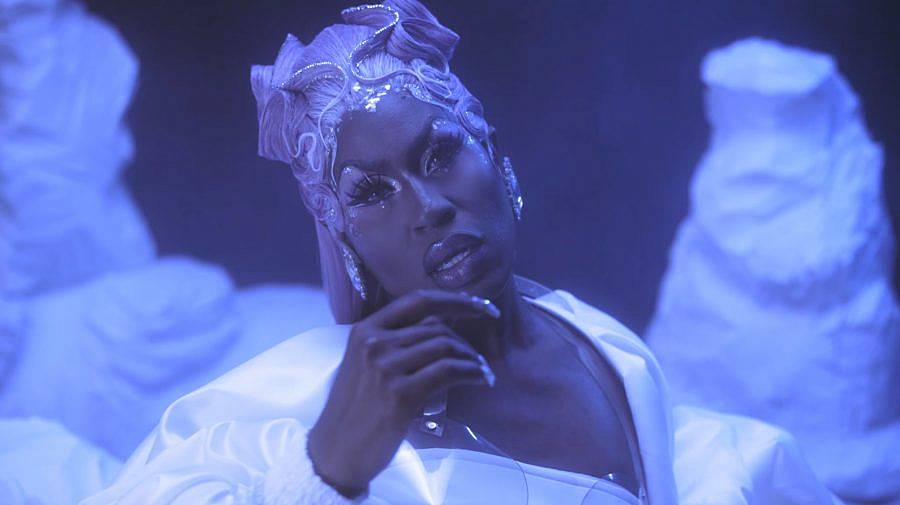 Image Description: This is a still of a drag queen all in blue. The lighting and background is blue with forms resembling icebergs in the background. She has an elaborate updo and long nails.