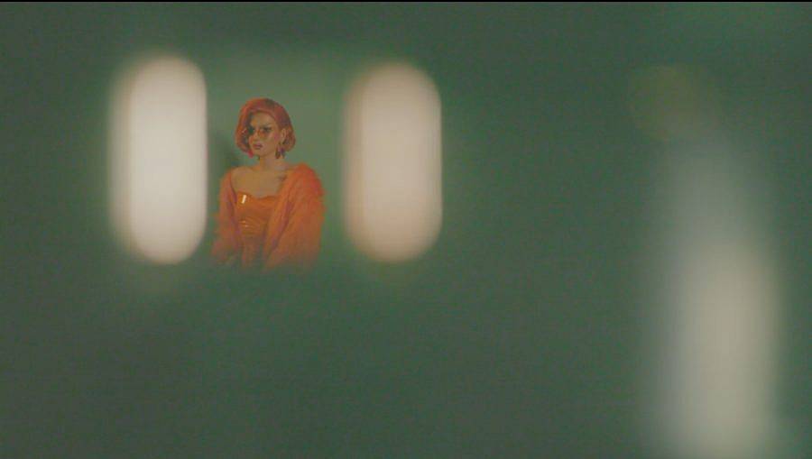 Image Description: This is a still of a drag queen sitting in an empty room. They are dressed all in bright orange with orange hair. All the space around her is blurry.