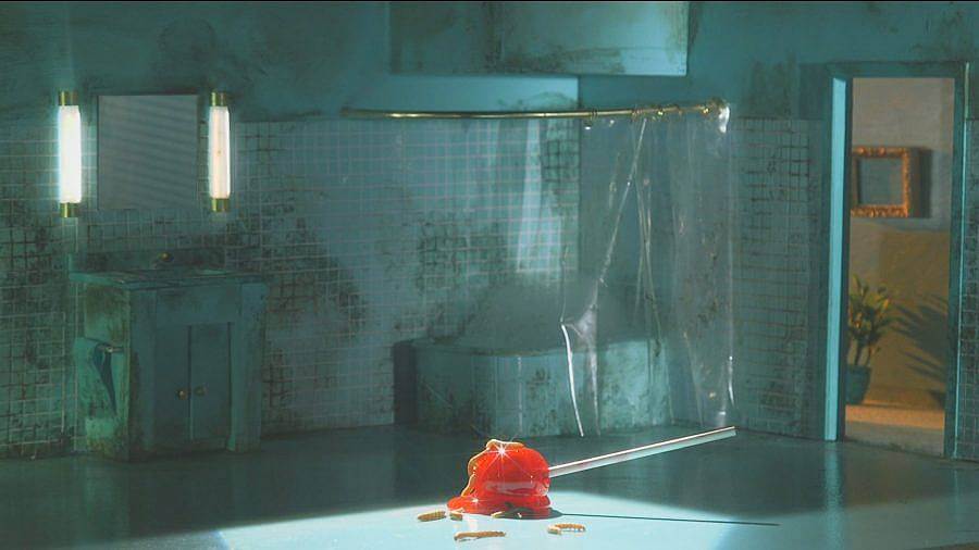 Image Description: This is a still showing a very dirty tiled bathroom. There is a clear shower curtain and a door looking into a hallway with plants. A giant red lollipop is melting in the center of the floor.