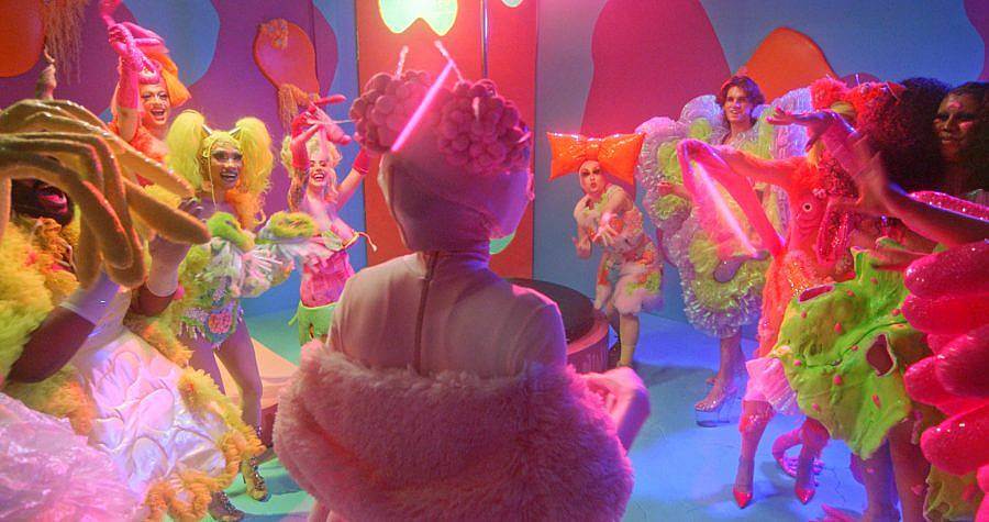 Image Description: This is a still with many people in elaborate colorful costumes with big hairstyles. The set is also multi-colored and a party appears to be happening.