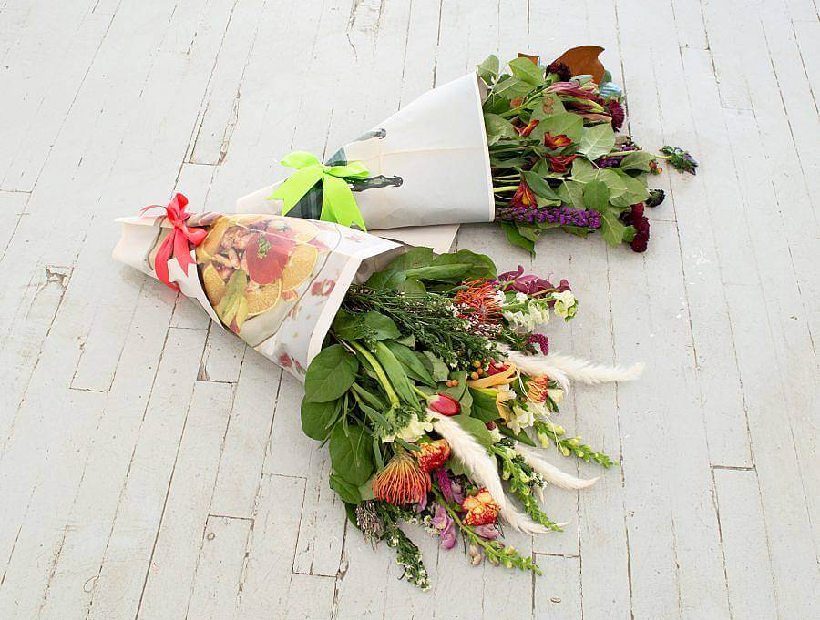 Image Description: There are two floral bouquets laying on the floor. Both bouquets are wrapped in photographs printed on newsprint. There are green and pink ribbons tied around them.