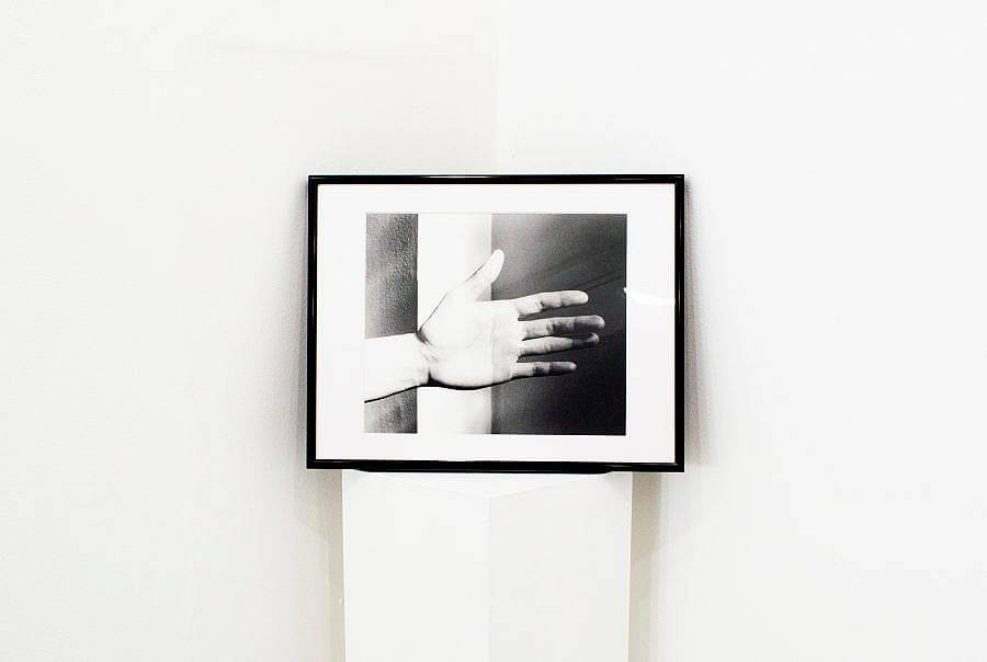 Image Description: This is a detail shot. This is a framed photograph of a hand sitting on a pedestal. The photograph is propped up against the corner of the wall. There is a beam of light moving down the center of the hand.