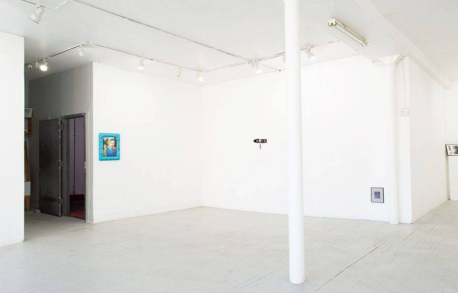 Image Description: This is an installation shot of the gallery. Close to the edge of the wall is a photograph of a clown in a blue satin frame. A pair of binoculars hangs from the ceiling in the center and there is a small black and white photo hung close to the floor.